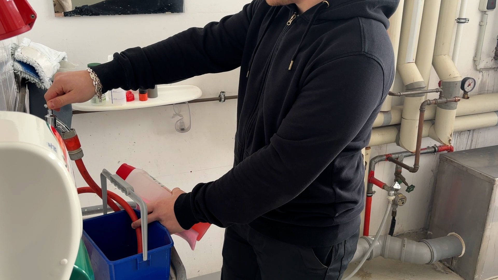 Eine Person steht in einem Waschraum oder Technikraum und füllt eine rosafarbene Flüssigkeit aus einer Flasche in einen blauen Behälter unter einem Wasserhahn.