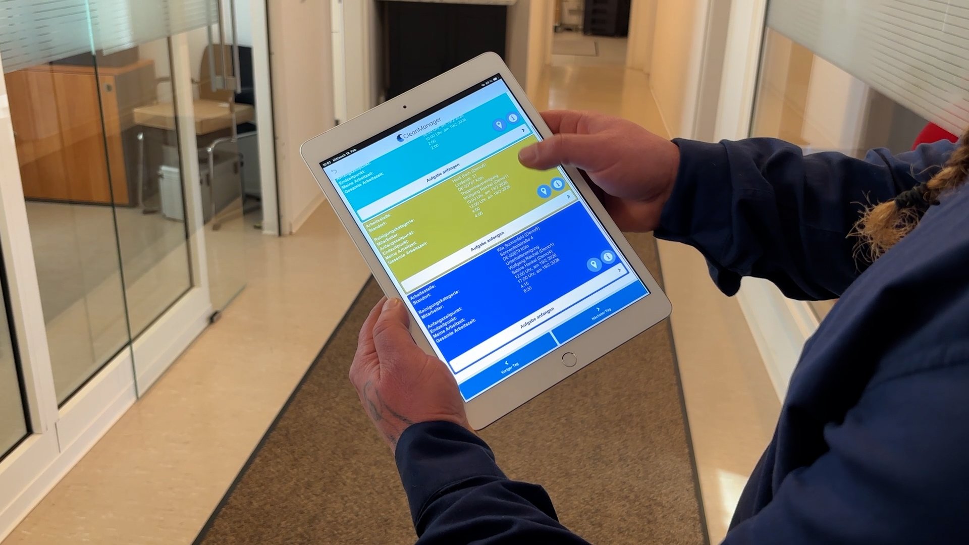 Eine Person hält ein Tablet und nutzt die CleanManager-App, die eine Oberfläche mit farblich strukturierten Bereichen zeigt.